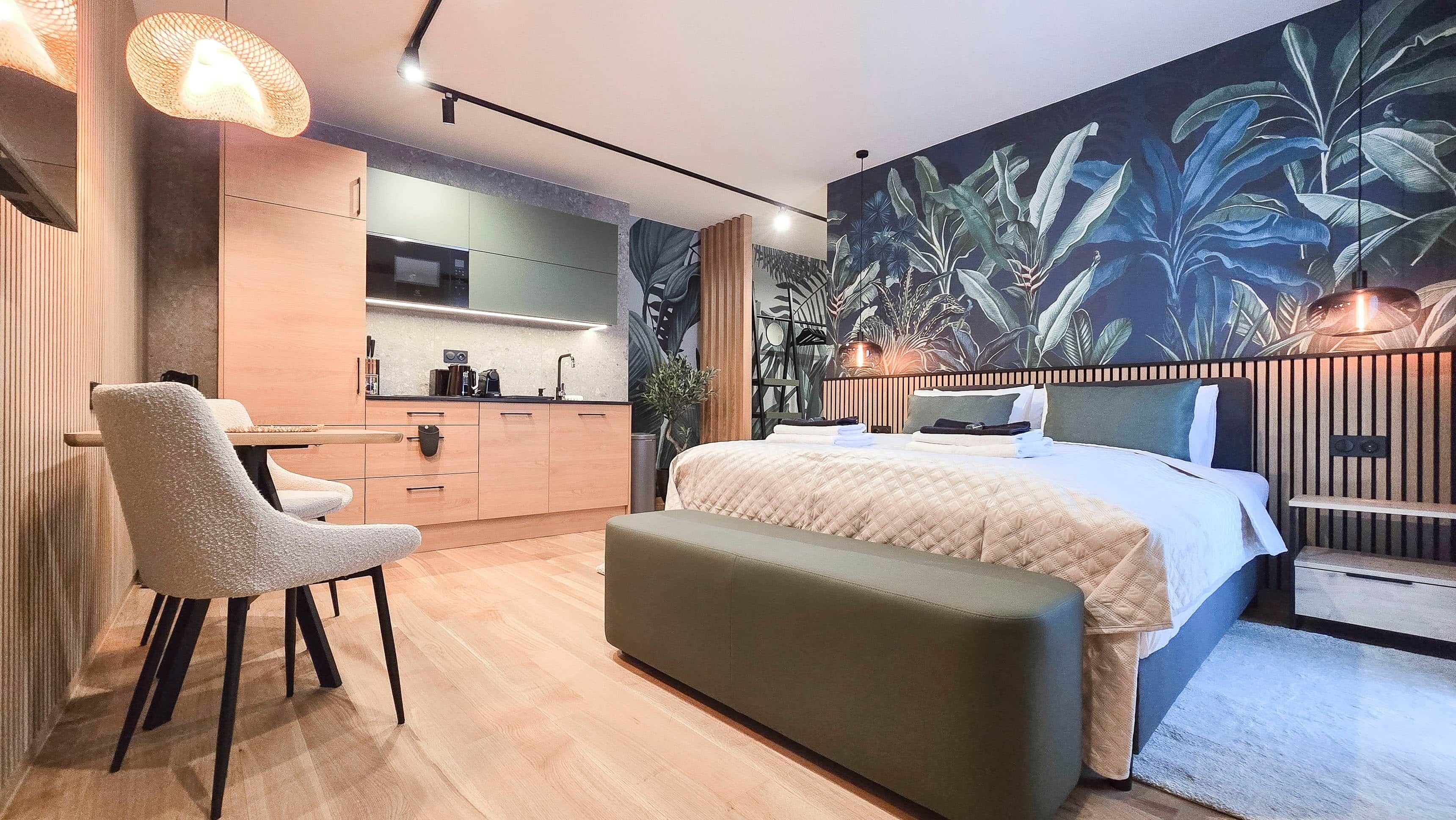 A modern bedroom with a large bed, a seating area with a chair, a small kitchenette, and a wall with a tropical plant mural.