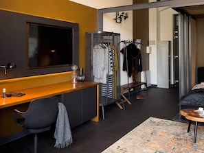 A modern hotel room with a large flat-screen TV, a wooden desk, a seating area with a patterned rug, and a walk-in closet with hanging clothes. - IMCT - Astoria Central Home 1 (Budapest)