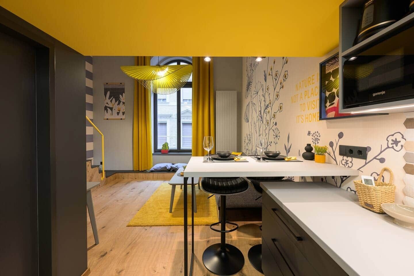 Een moderne keuken met een wit aanrecht, zwarte barkrukken, een geel vloerkleed en een raam met gele gordijnen.