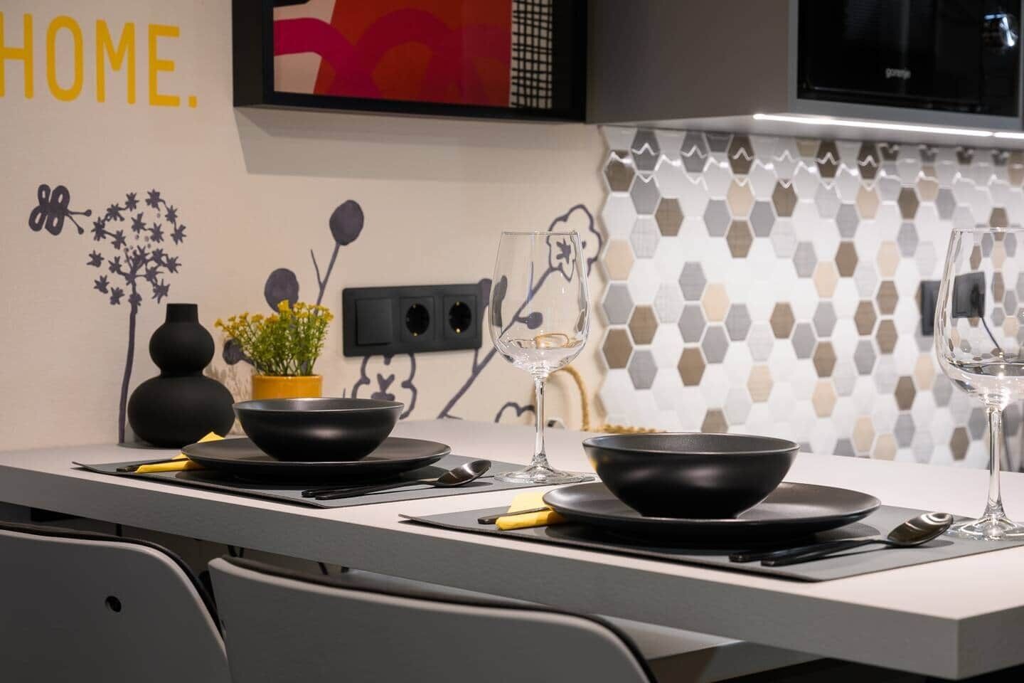 A modern kitchen with a dining table set for two, featuring black bowls, plates, and cutlery.