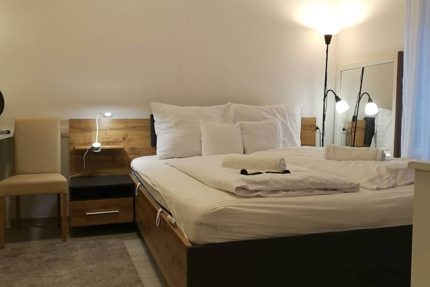 A neatly made bed with white linens, a bedside lamp, a wooden nightstand, and a chair.
