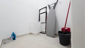 A room with a white wall, a black ironing board, a white folding chair, a black bucket, and a red mop. - Central Vibe Apartments 1 (Budapest)