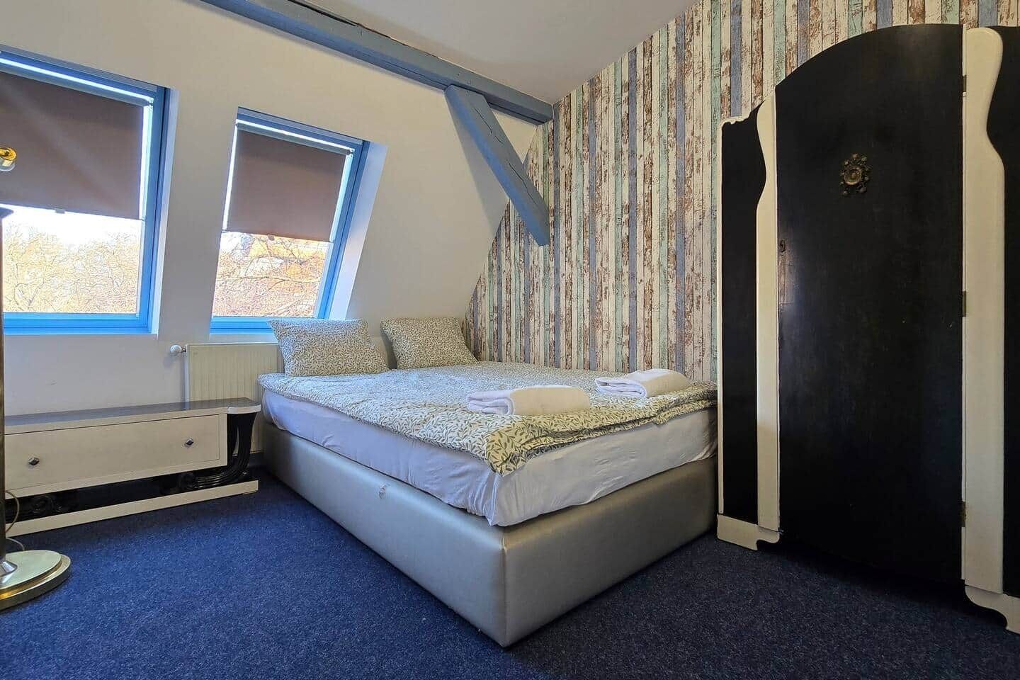 A bedroom with a bed, two windows, a radiator, a wardrobe, and a desk.