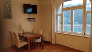 A dining area with a table and chairs, a television mounted on the wall, and a window with a view of a body of water and hills. - IMCT - Danube View Apartment (Budapest)