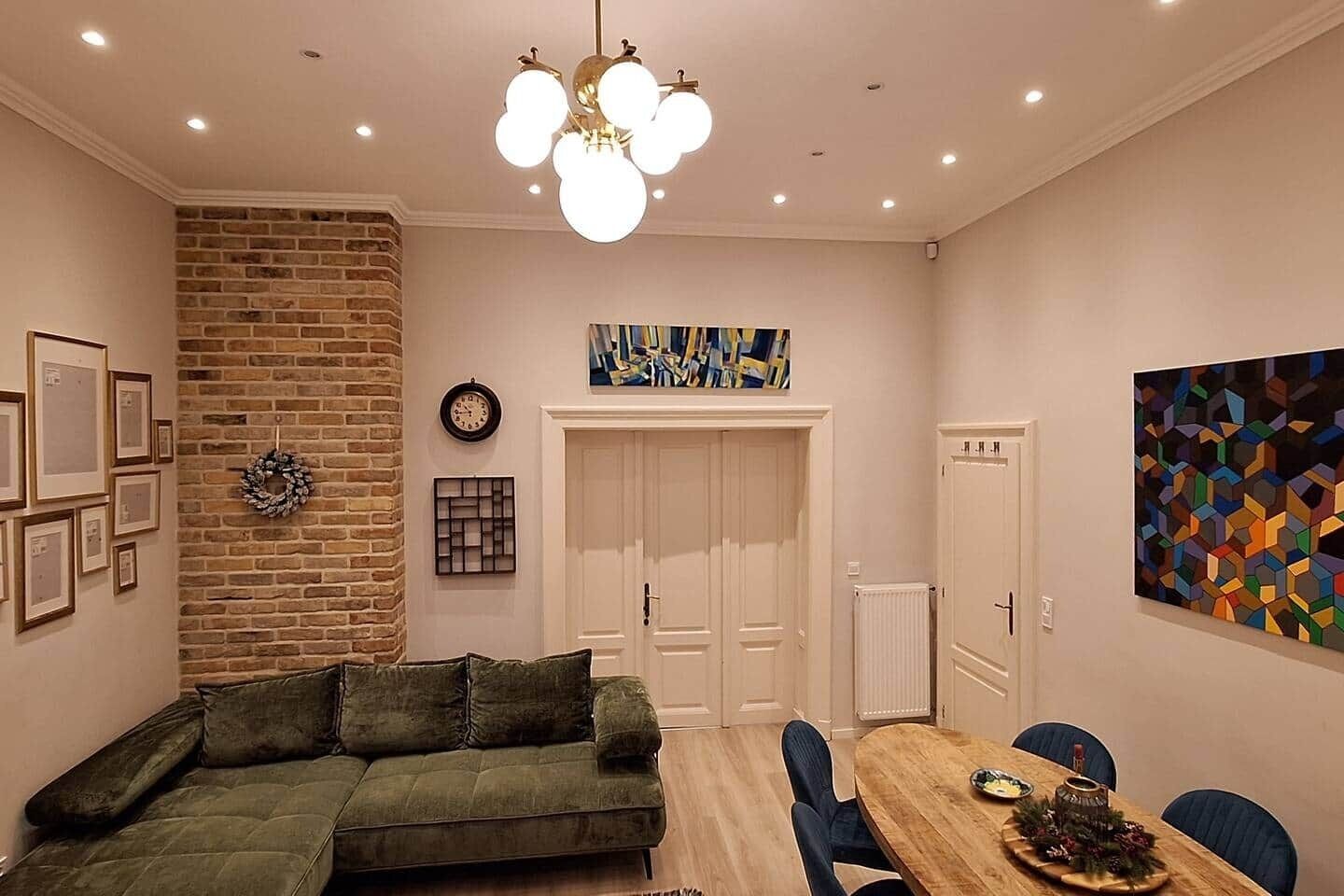 A dining area with a wooden table, blue chairs, a green sofa, a brick wall, and a modern light fixture.