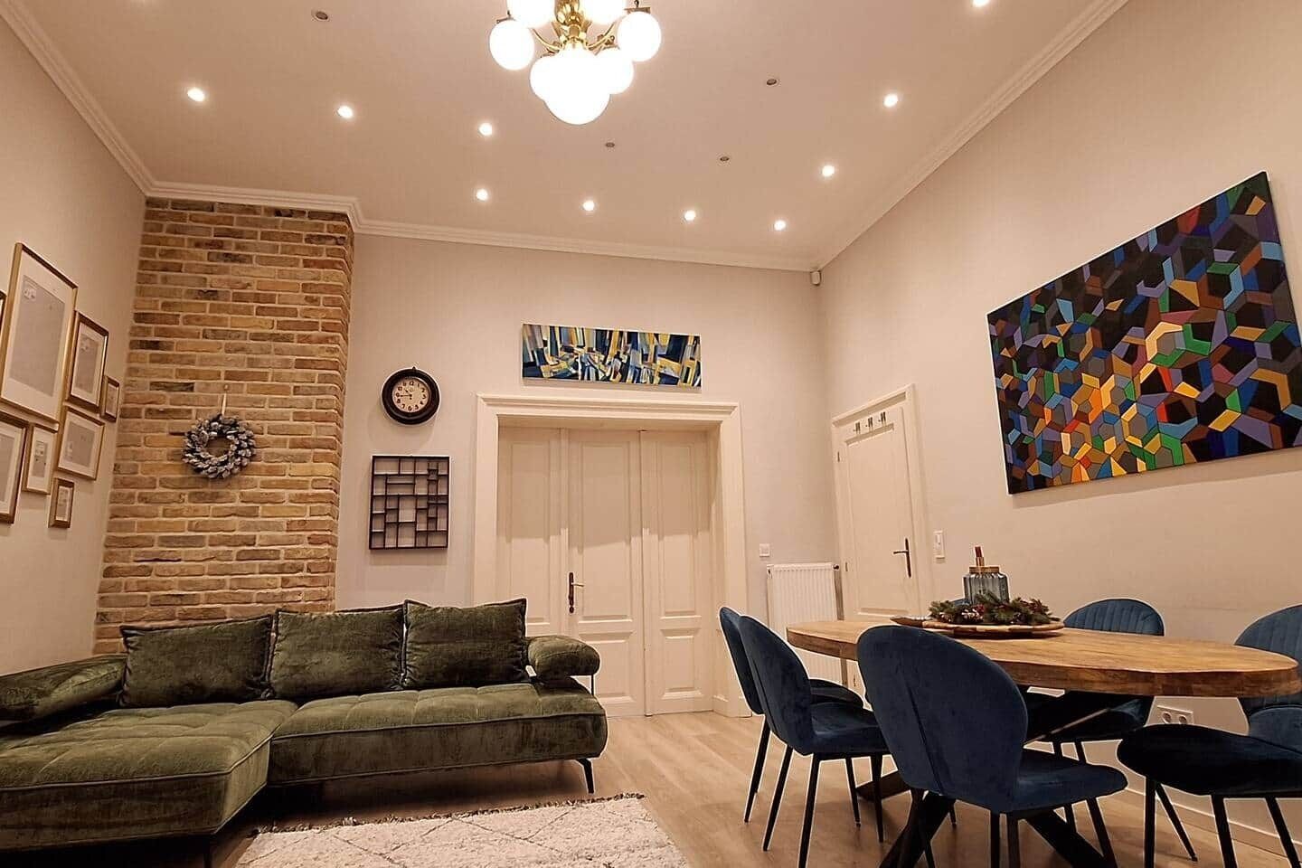 A living room with a green sofa, a dining table with chairs, a brick wall, and a colorful abstract painting.