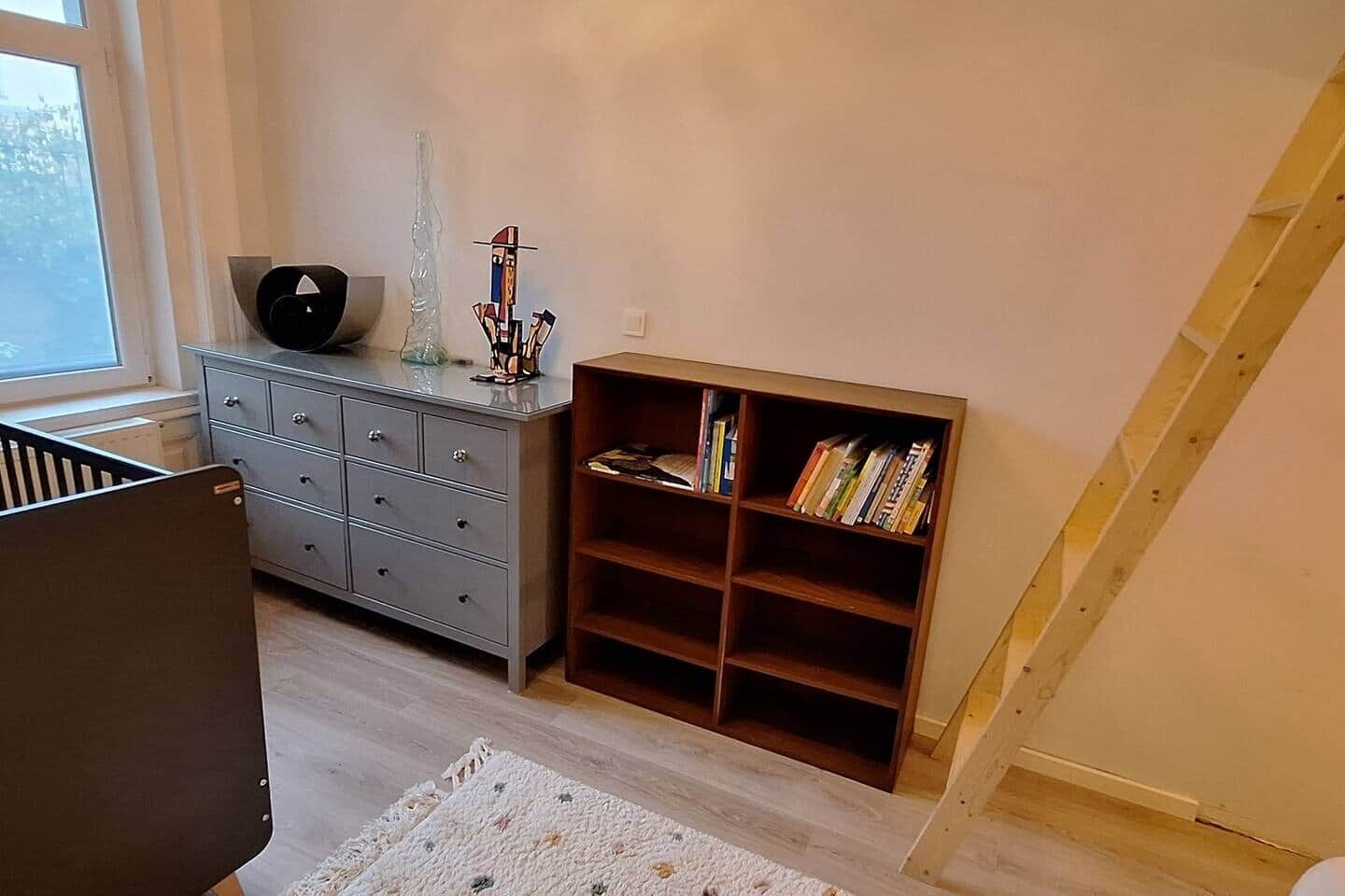 A room with a crib, a grey dresser, a bookshelf, and a window.