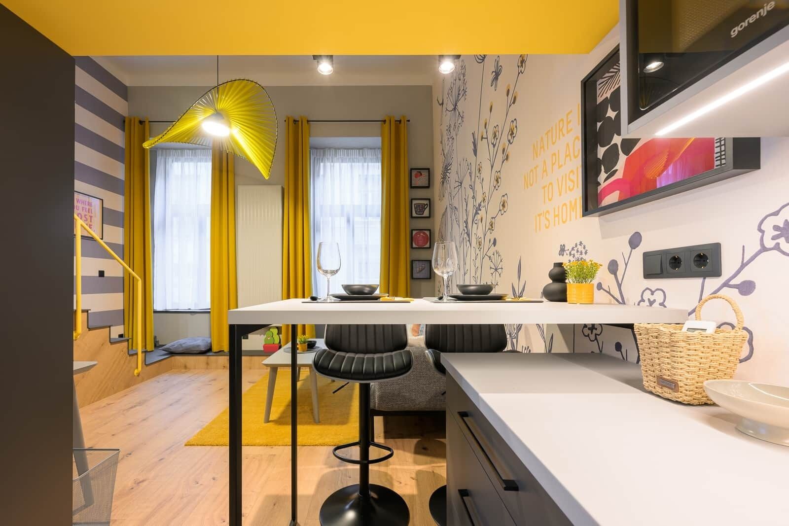 A modern kitchen with a white countertop, two bar stools, a yellow pendant light, and a wall with floral wallpaper.