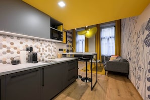 A compact kitchen with a microwave, toaster, and coffee machine. There's a dining area with a table and chairs, and a sofa in the background. - IMCT - Dockhouse Soho 3 (Budapest)