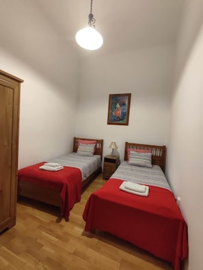 A small, single-room accommodation with two wooden beds, each with a red bedspread and pillows, a wooden wardrobe, a framed picture on the wall, and a hanging light fixture. - IMCT - Elegance in the Heart (Budapest)