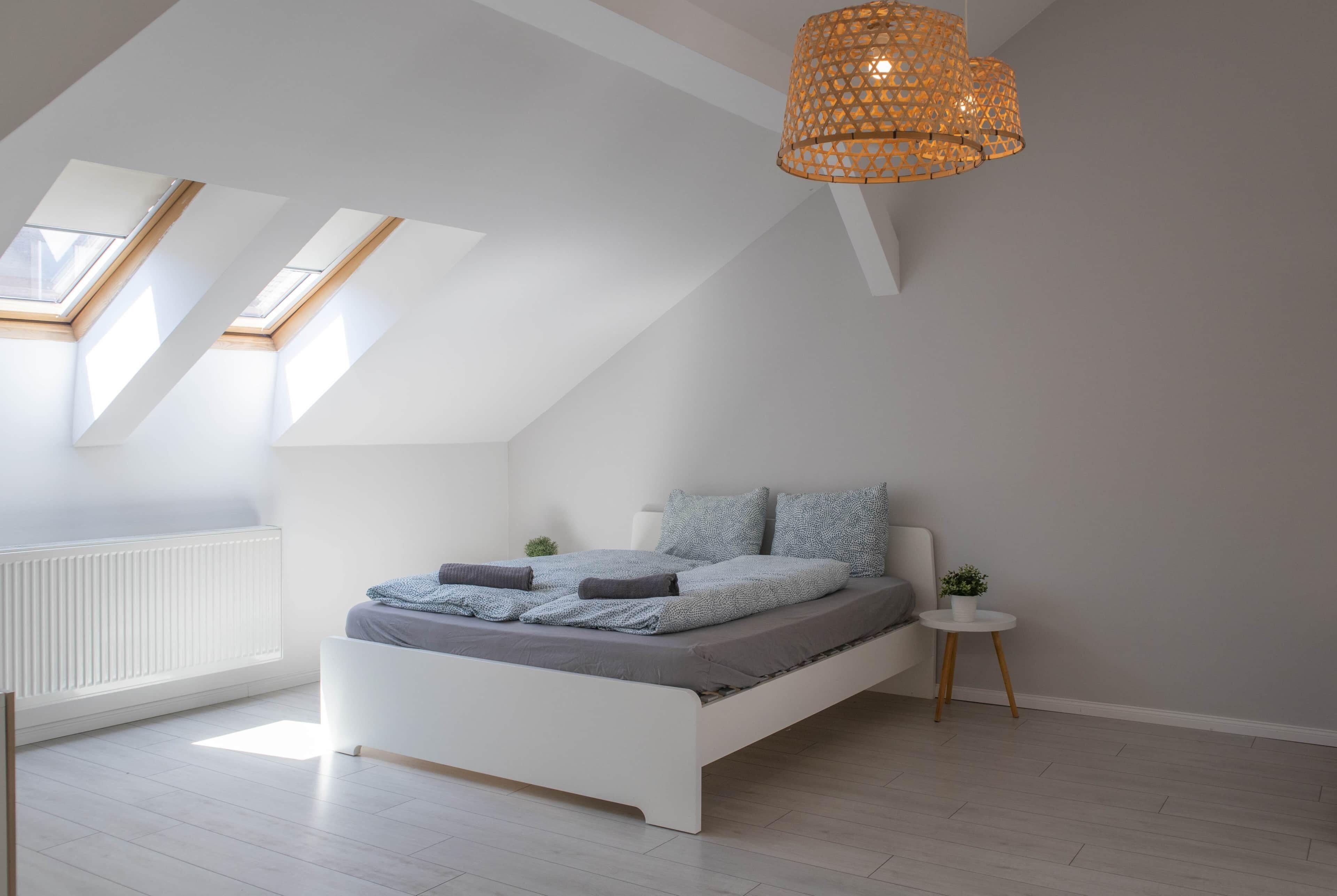 A modern bedroom with a sloped ceiling, skylights, a bed with grey bedding, a small bedside table with a plant, and a hanging light fixture.