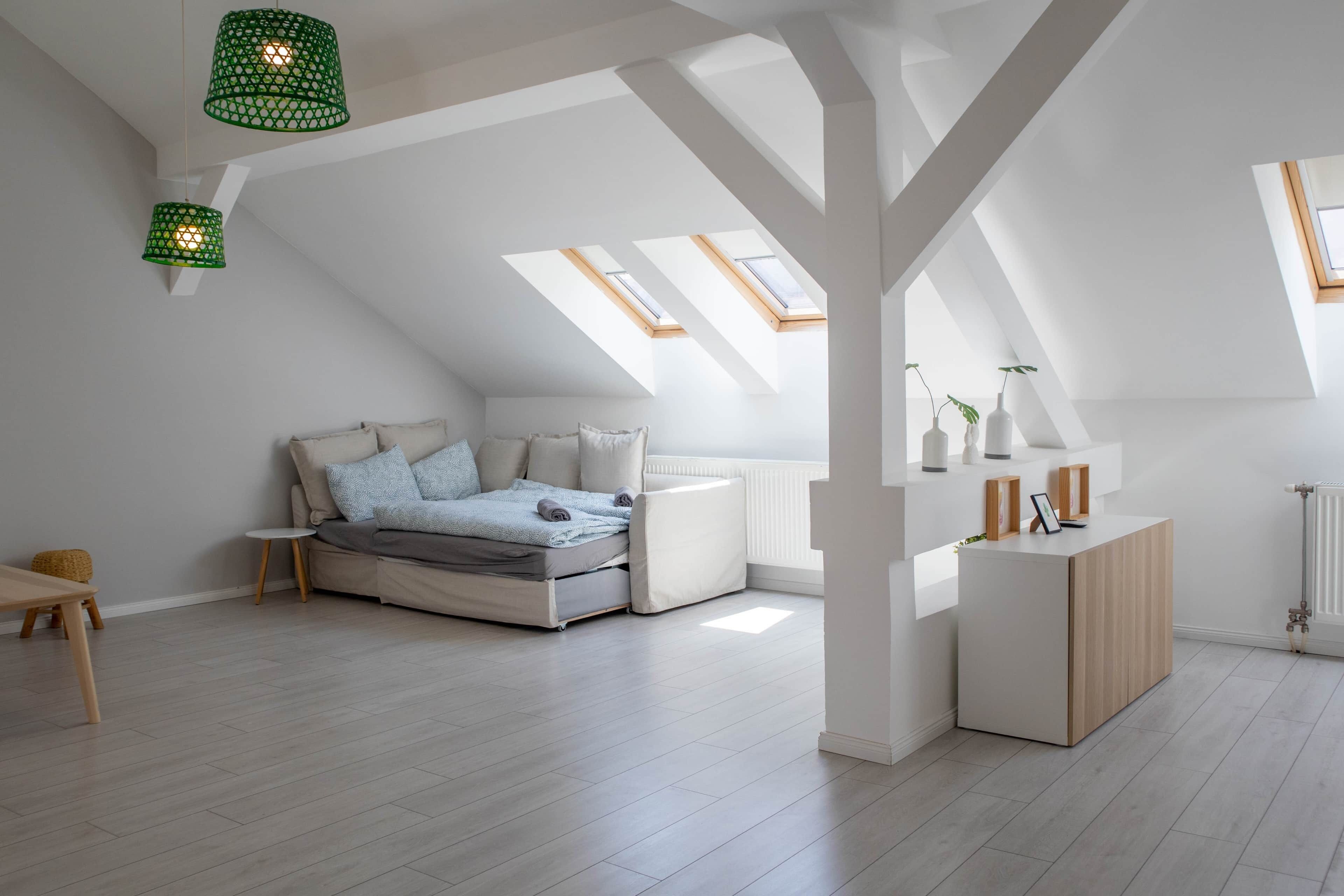 A modern loft bedroom with a sloped ceiling, skylights, a bed with a grey mattress and patterned pillows, a small bedside table, and a wooden nightstand with decorative items.