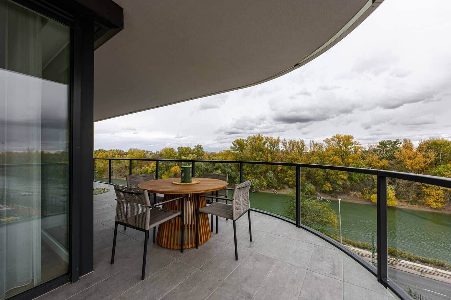 A balcony with a round table and chairs, overlooking a river and autumn foliage.
