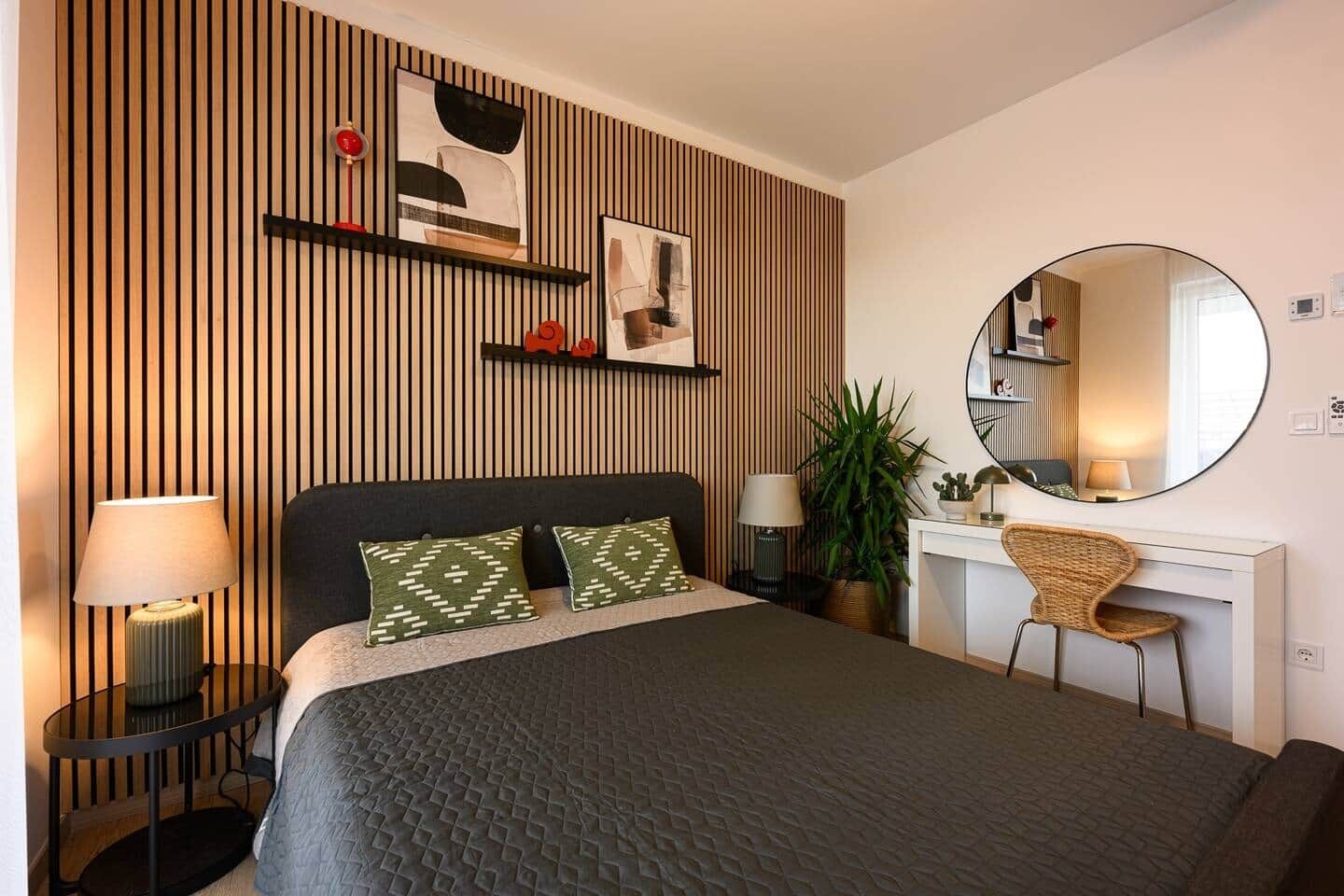 A hotel room with a bed, bedside tables, a desk, a chair, a round mirror, and a potted plant.