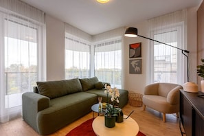 A modern living room with a green sofa, a beige armchair, and a coffee table with a plant. There are large windows with sheer curtains, a floor lamp, and artwork on the wall. - IMCT - OpenSky Terrace with free parking (Budapest)