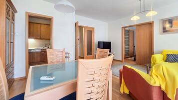 Uma sala de estar moderna com uma mesa de jantar de vidro, cadeiras de madeira, um sofá amarelo e uma TV.