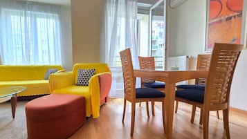 Ein Wohnzimmer mit einem gelben Sofa, einem roten Hocker, einem Holzesstisch mit blauen Stühlen und transparenten Vorhängen.