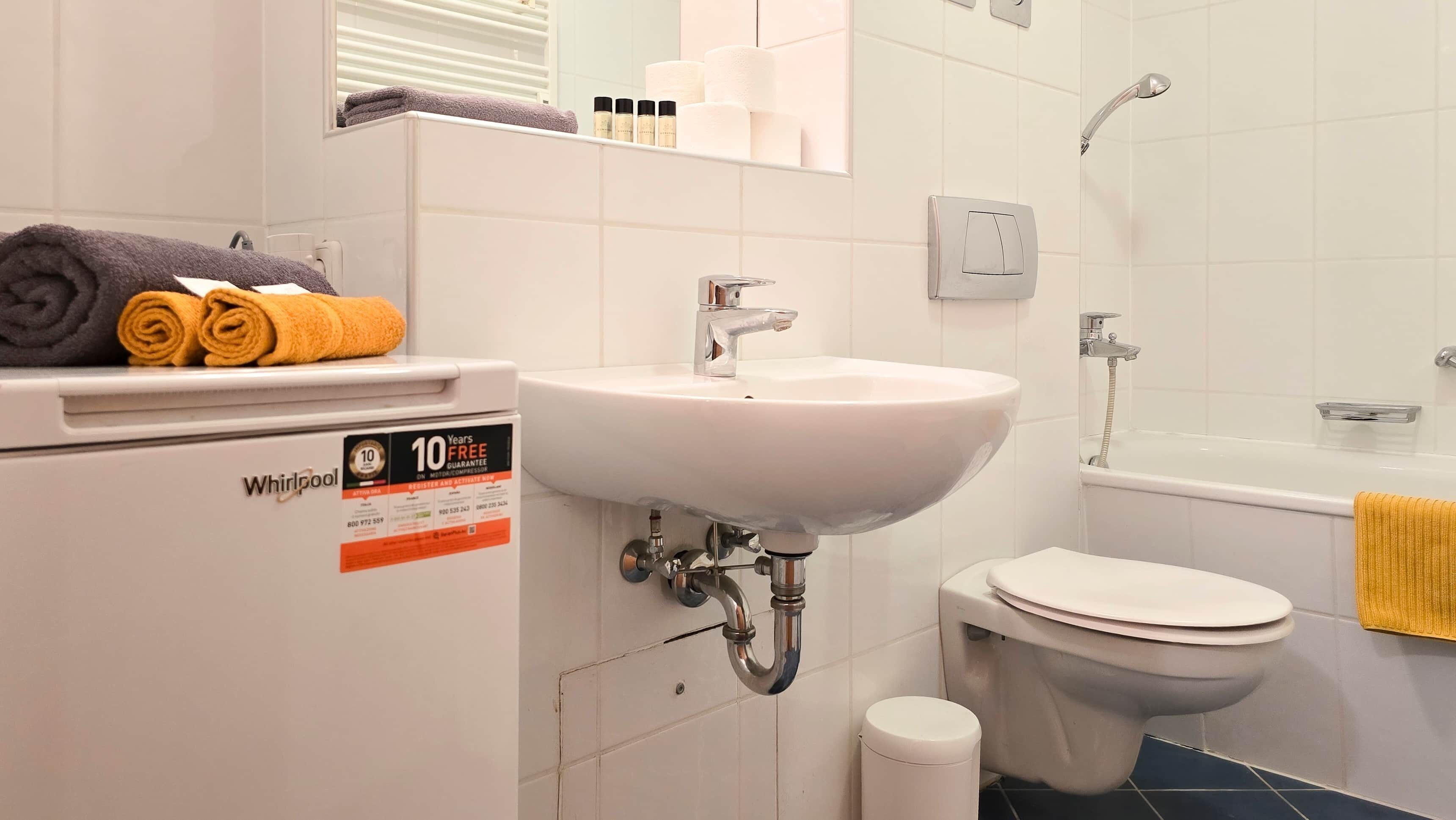Une salle de bain équipée d’un lavabo, de toilettes et d’une douche. Des serviettes sont posées sur une étagère, et un panneau indique une garantie de 10 ans.