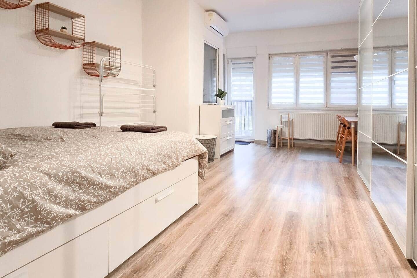 Una camera da letto moderna con un letto grande, una cassettiera e una scrivania con una sedia. Si vede una finestra con persiane e un termosifone.