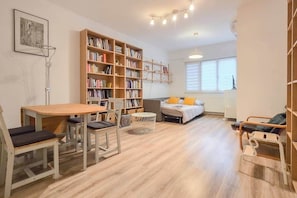 Un soggiorno accogliente con pavimento in legno, un tavolo rotondo con sedie, una libreria, un letto con cuscini e un lampadario.