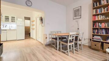 Un espacio habitable compacto con kitchenette, una zona de comedor con mesa y sillas de madera, y una estantería llena de libros.