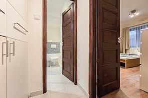 A hotel room with a wooden door, a bed, a window with curtains, and a bathroom with a toilet and sink. - IMCT - New City Life Apartment (Budapest)