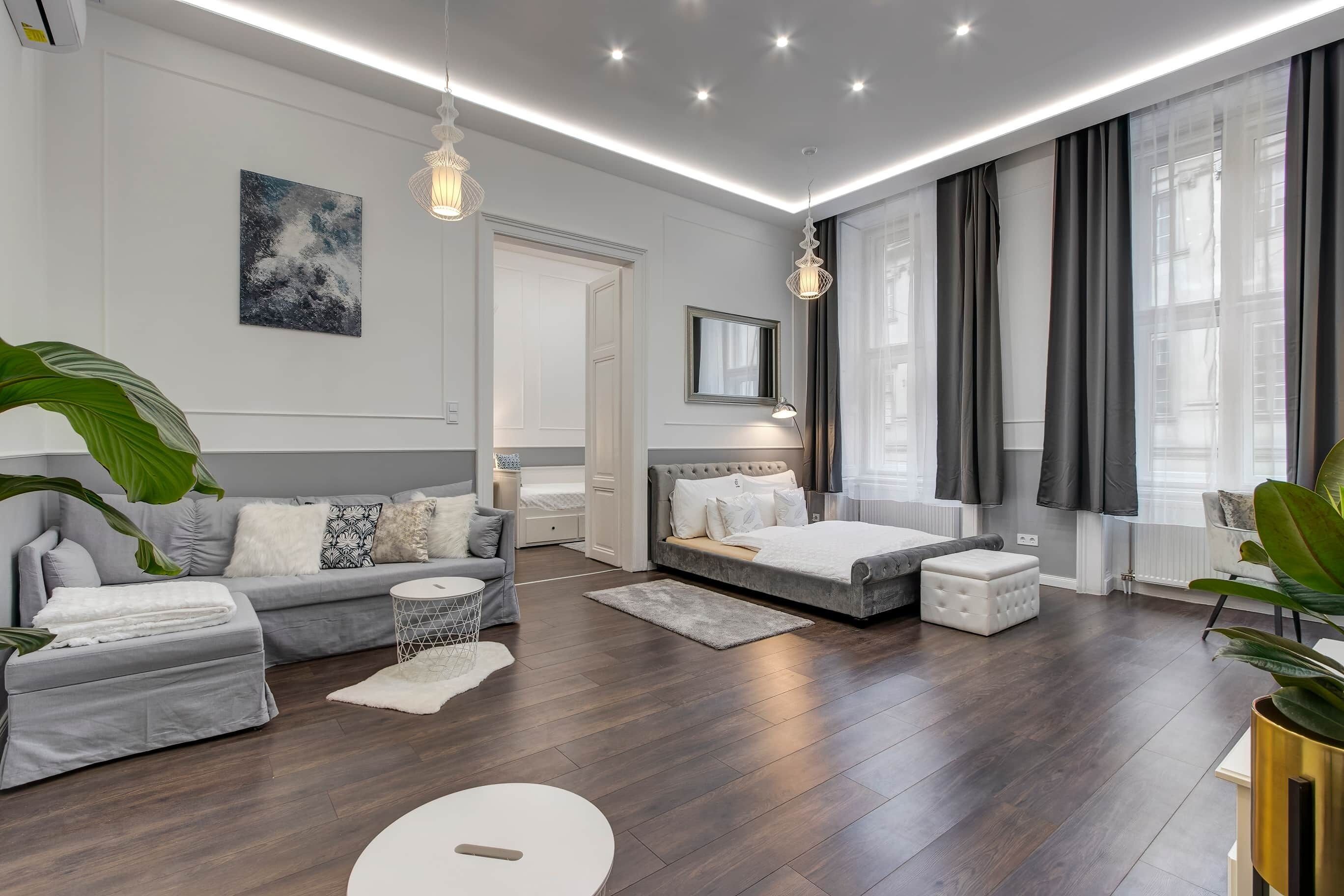 A modern bedroom with a grey sofa, a bed, a round white table, and a large window with curtains.