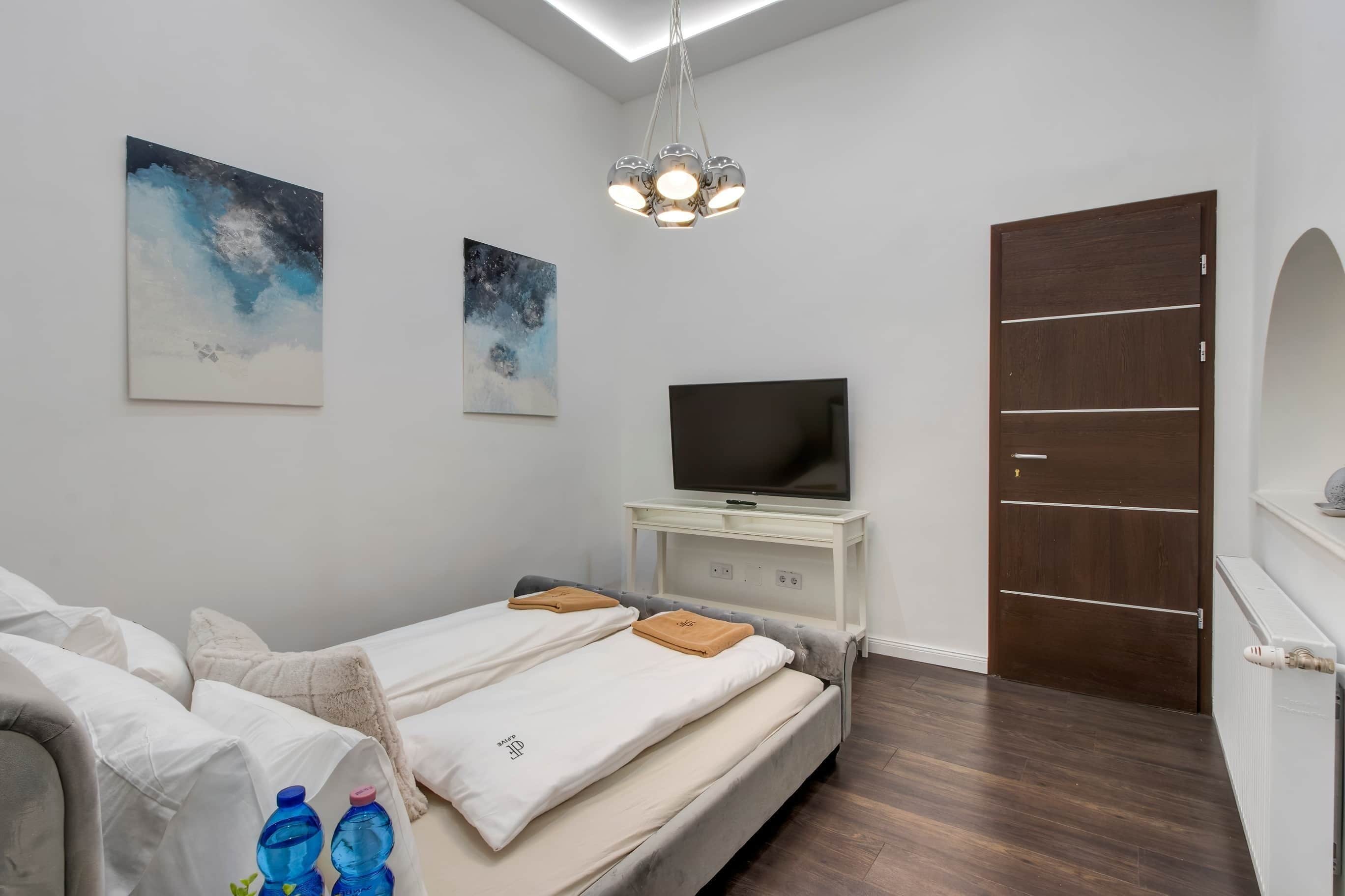 A bedroom with a bed, a TV on a stand, two paintings, a wooden door, and a ceiling light fixture.