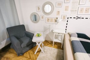 A room with a grey sofa, a white side table with a bowl of fruit, a white desk with a lamp, and a bed with a striped blanket. - IMCT - Tailor Apartment in the Center (Budapest)