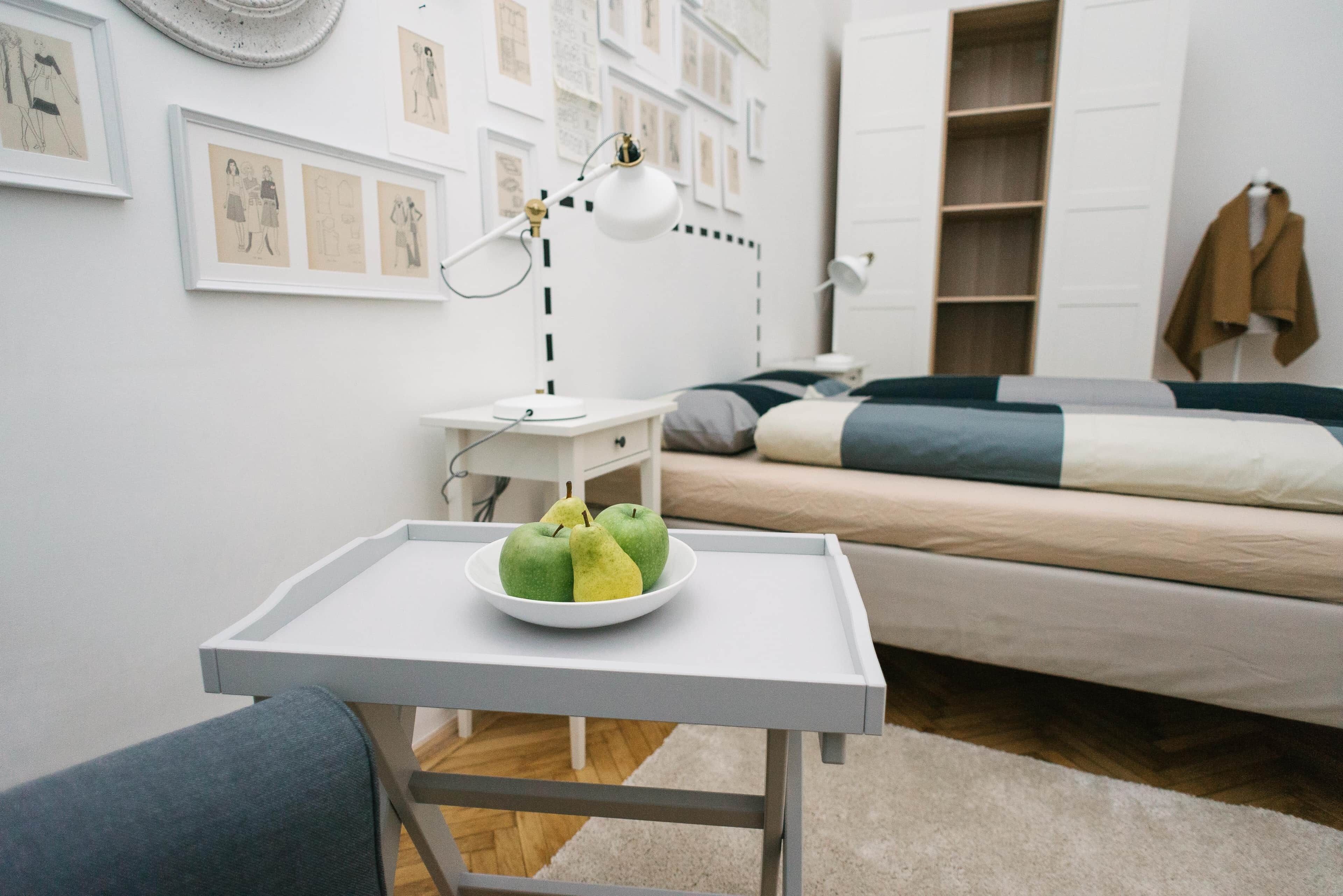 A bedroom with a bed, bedside table with a lamp, a wall with framed sketches, and a tray with apples and pears.