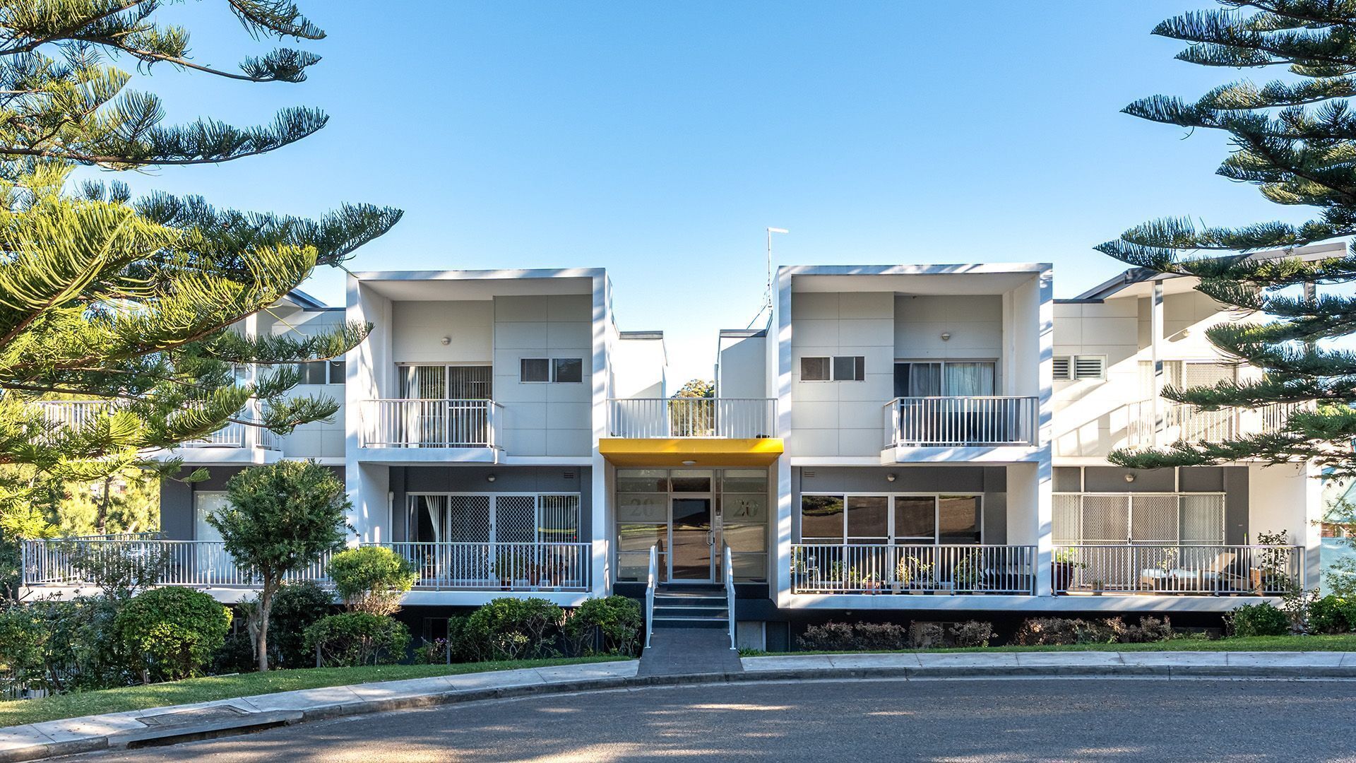 A modern two-story building with balconies, a central entrance, and a clear sky.