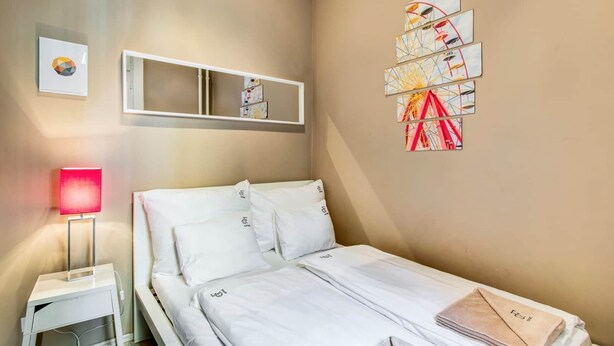 Ett hotellrum med en säng, ett sängbord med en lampa, en vägghylla med konstverk och ett stort fönster.
