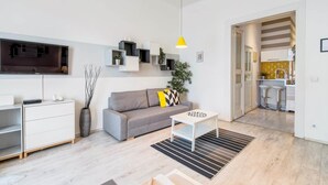 Ett modernt vardagsrum med en grå soffa, ett soffbord, en tv som är monterad på väggen och ett kök som syns genom en öppen dörr.