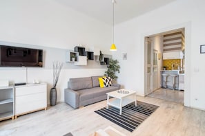 A modern living room with a grey sofa, a coffee table, a TV mounted on the wall, and a kitchen visible through an open door. - IMCT - Panorama Apartment Synagogue (Budapest)