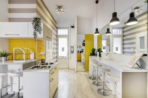 A modern kitchen with a central island, bar stools, pendant lighting, and a yellow accent wall. - IMCT - Panorama Apartment Synagogue (Budapest)