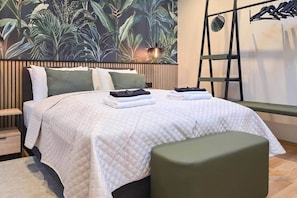 A bedroom with a bed, pillows, a bench, a hanging light, and a wall with a plant mural. - IMCT - Urban Jungle Apartments 09 (Budapest)
