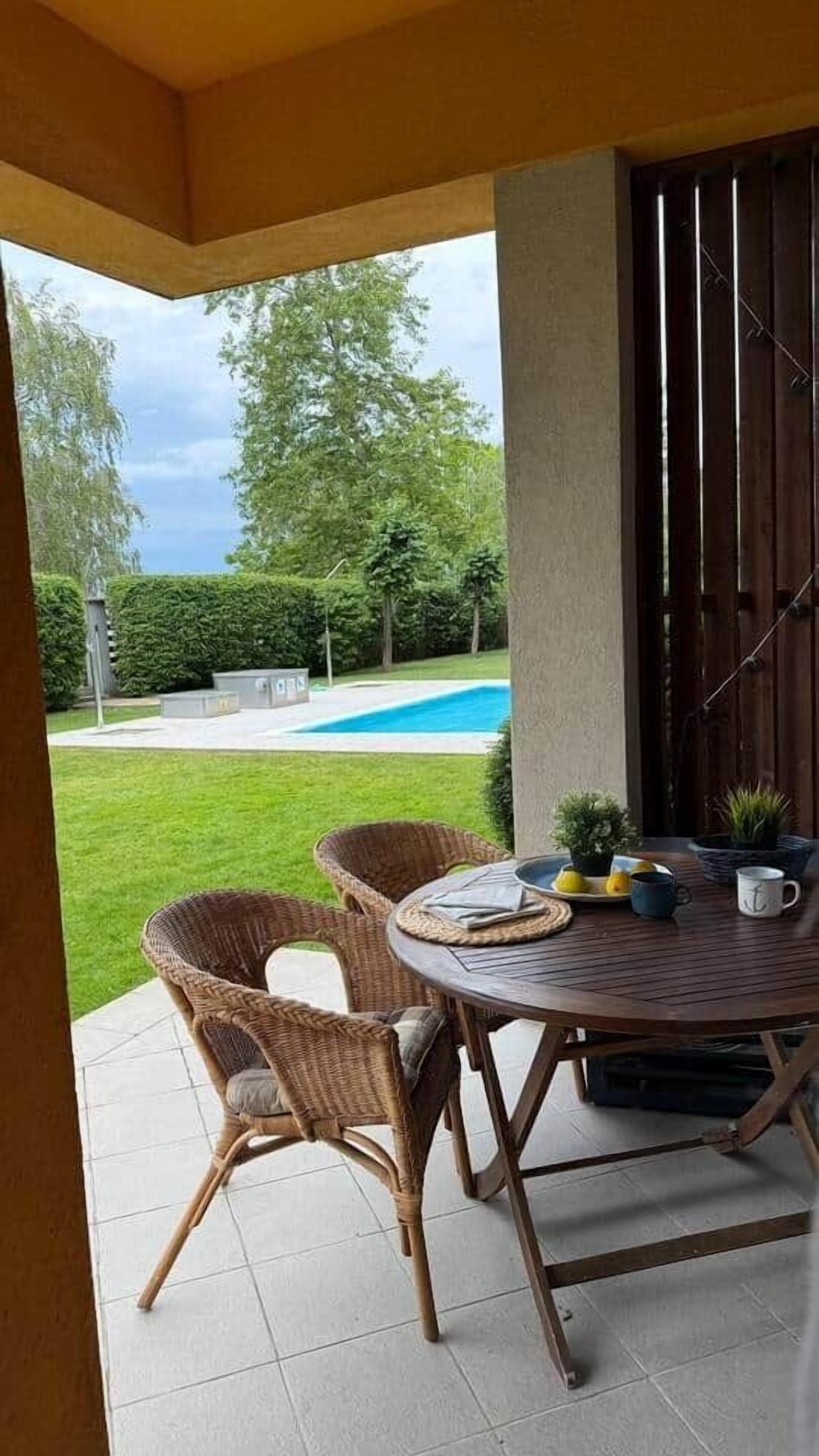 An outdoor patio with wicker chairs and a table, overlooking a swimming pool and a well-maintained lawn.