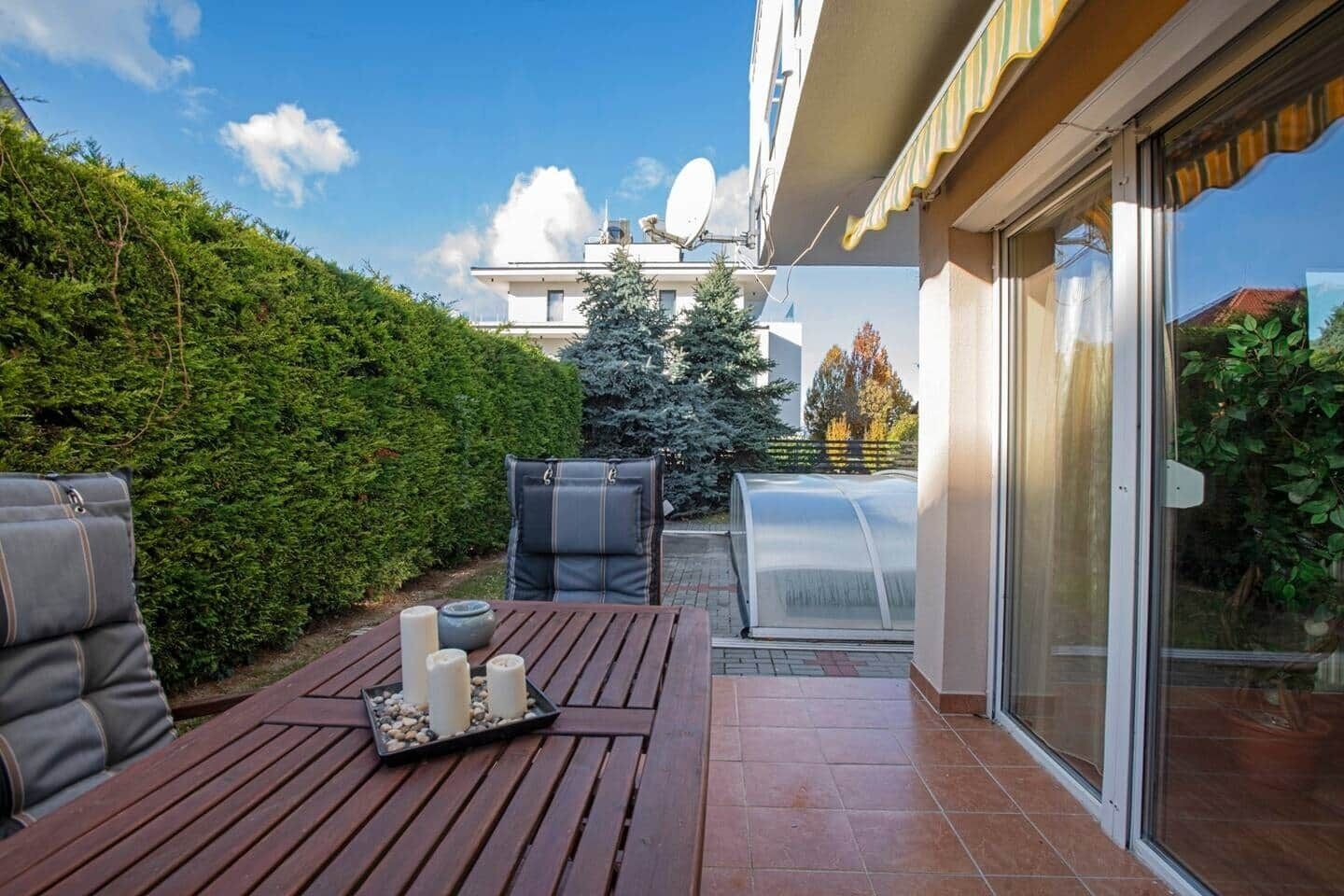A patio with a wooden table, cushions, and a greenhouse.