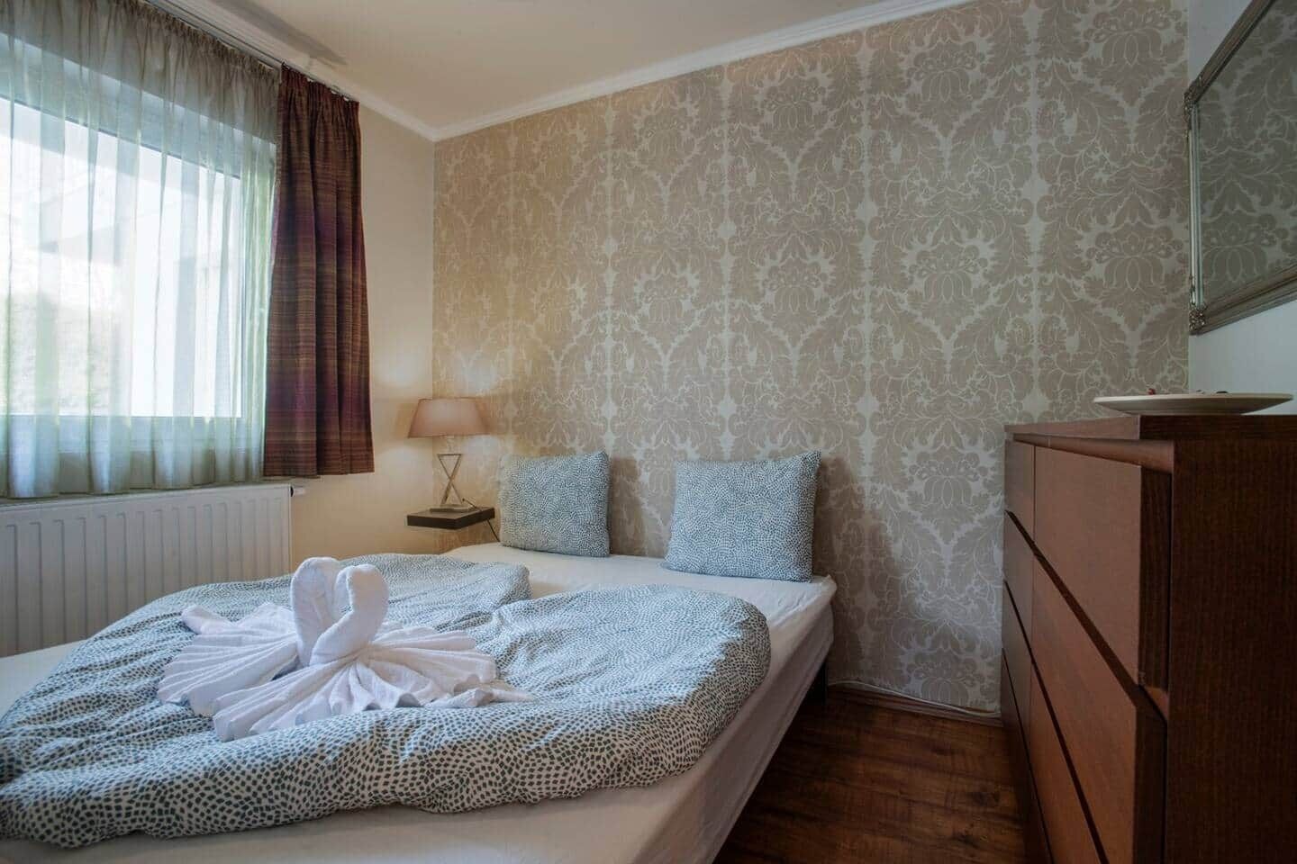 A hotel room with a bed, a dresser, a nightstand with a lamp, and patterned wallpaper.