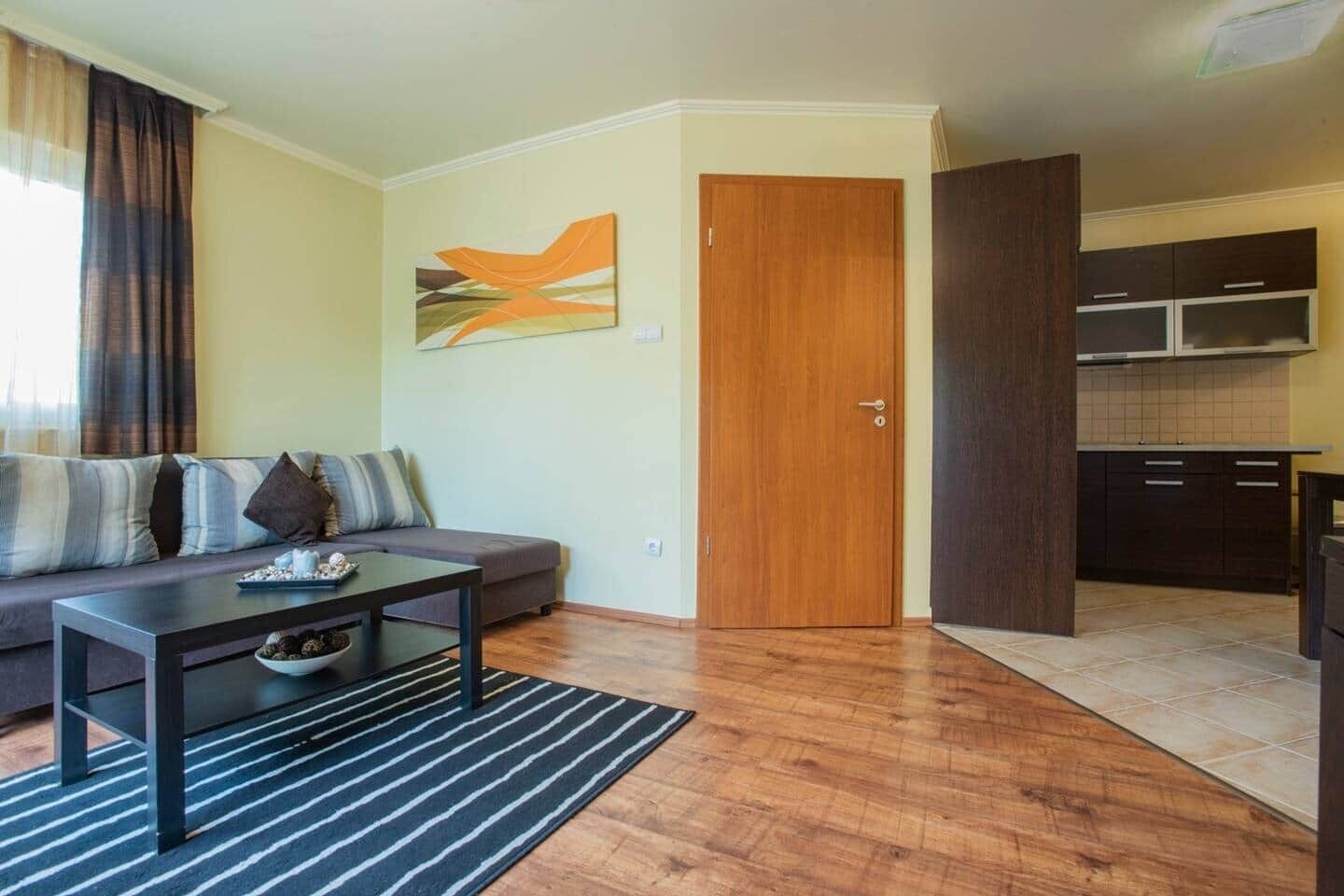 A living room with a sofa, coffee table, and a rug. There is a wooden door and a kitchen visible in the background.
