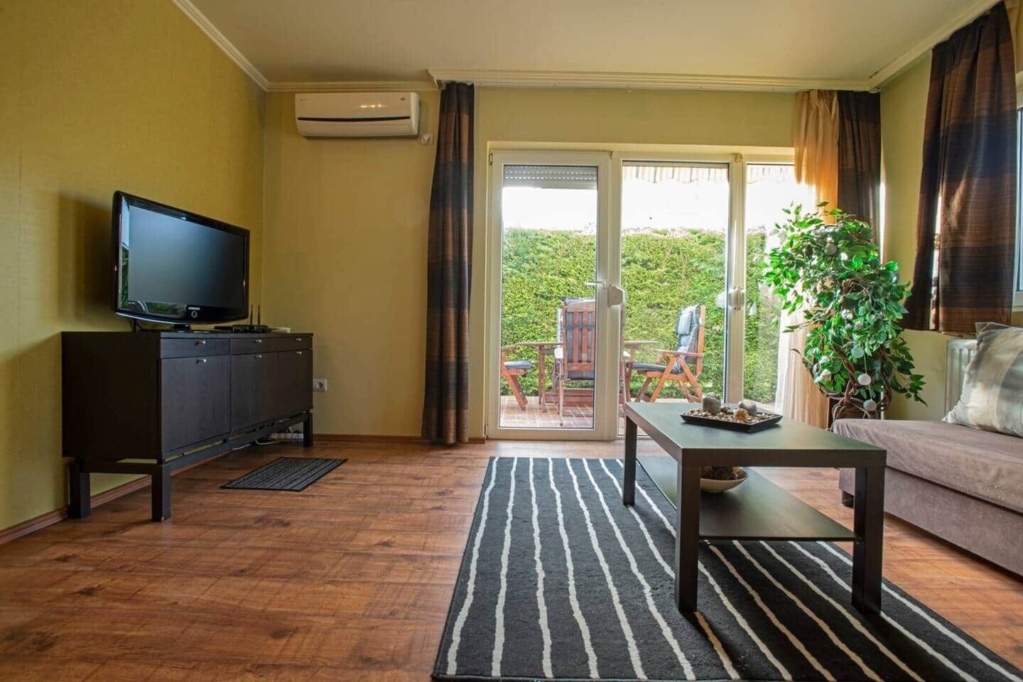 A living room with a flat-screen TV, a dark wooden cabinet, a coffee table, a sofa, a potted plant, and a sliding glass door leading to an outdoor seating area.