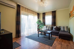 Sala de estar con sofá, una mesita de centro y una planta. Hay un radiador en la pared y una ventana con cortinas.