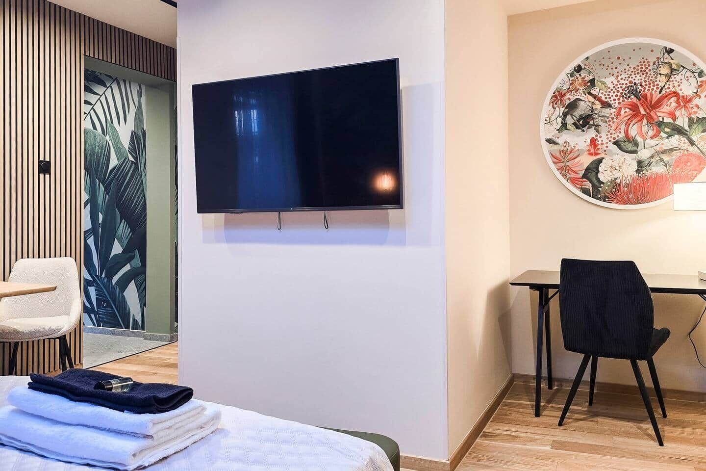 A hotel room with a bed, a desk, a chair, a television mounted on the wall, and a large floral artwork on the wall.