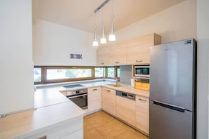 Cozinha moderna com geladeira de aço inoxidável, micro-ondas e forno embutido. Há luminárias pendentes sobre a bancada e uma janela que oferece iluminação natural.