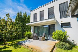 Ein modernes, zweistöckiges Haus mit überdachter Terrasse, einer großen Schiebetür und einer kleinen Sitzgruppe mit zwei Stühlen.