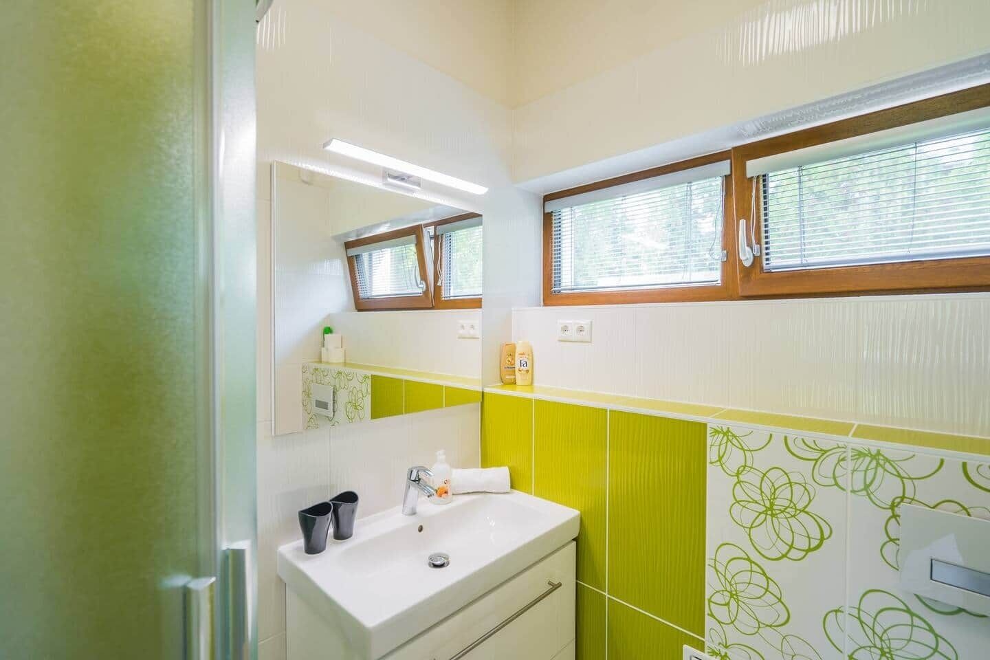 Ein Badezimmer mit einem weißen Waschbecken, grünen Fliesen mit Muster, einem Spiegel und einem Fenster mit Jalousien.