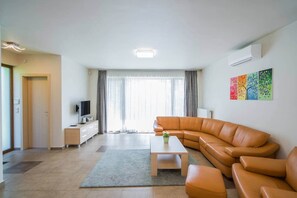 Ein modernes Wohnzimmer mit einem Ecksofa, einem Flachbildfernseher, einem Couchtisch und einem großen Fenster mit Vorhängen.