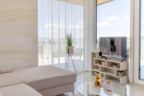 A modern living room with a sofa, a glass coffee table, and a TV on a stand. There is a balcony with a view of the city. - LB - LelleMarine Apartments A401 (Balatonlelle)