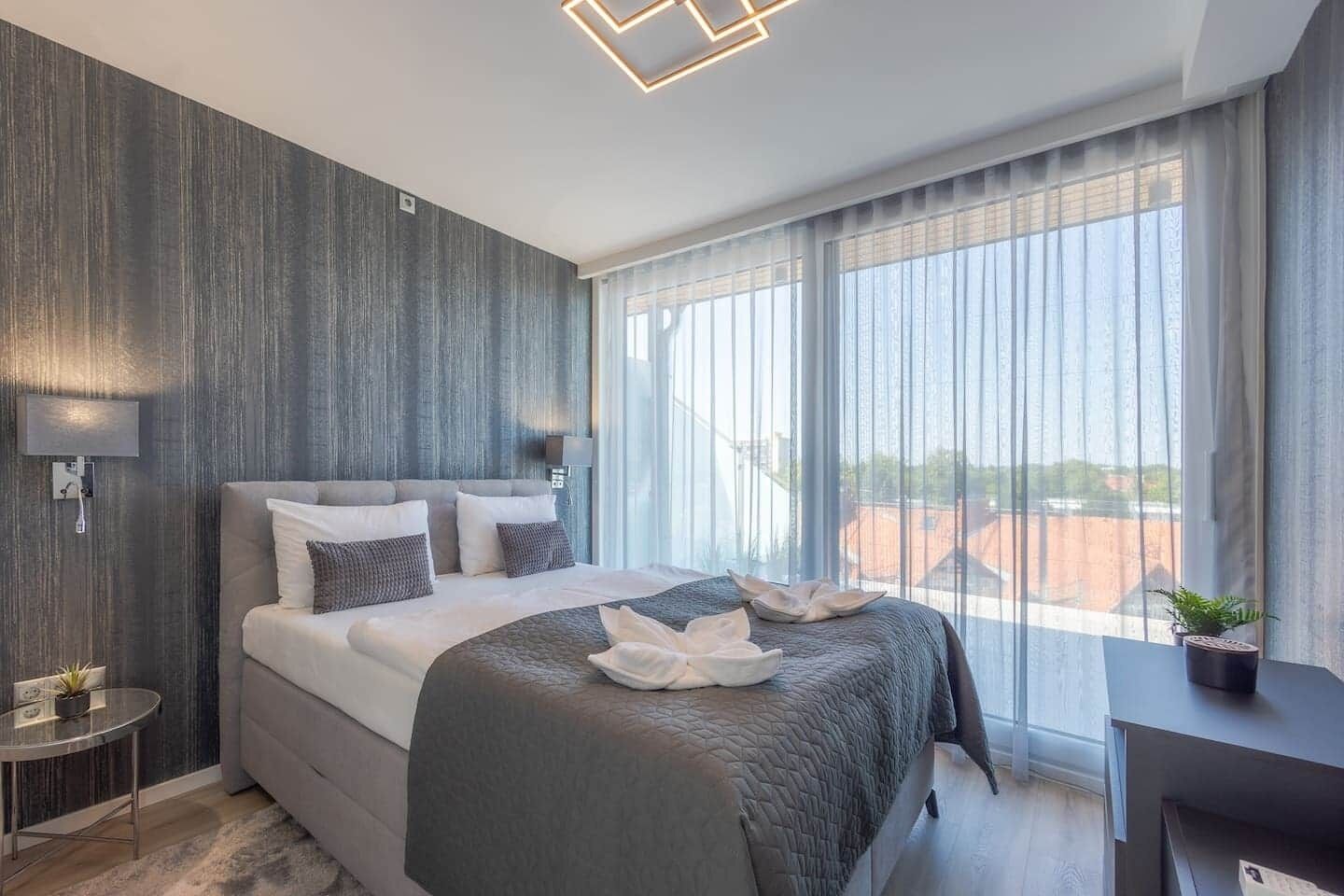 Una habitación de hotel moderna con una cama grande, una mesita de noche, un escritorio y vistas a la ciudad a través de amplias ventanas.
