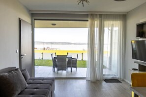 A modern living room with a sofa, a flat-screen TV, a balcony with a table and chairs, and a view of a body of water. - LB - LelleMarine Apartments A002 (Balatonlelle)